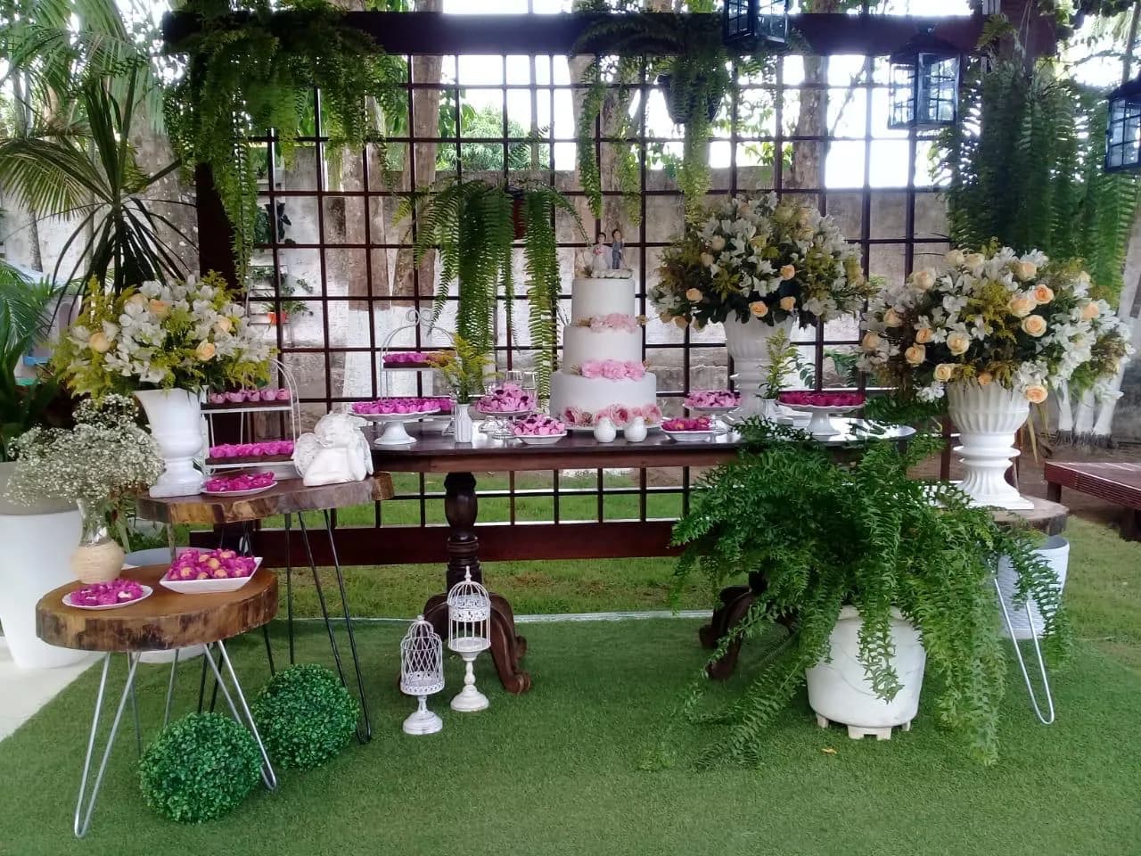 Mesa de doces montada para casamento, com painel de flores e samambaias