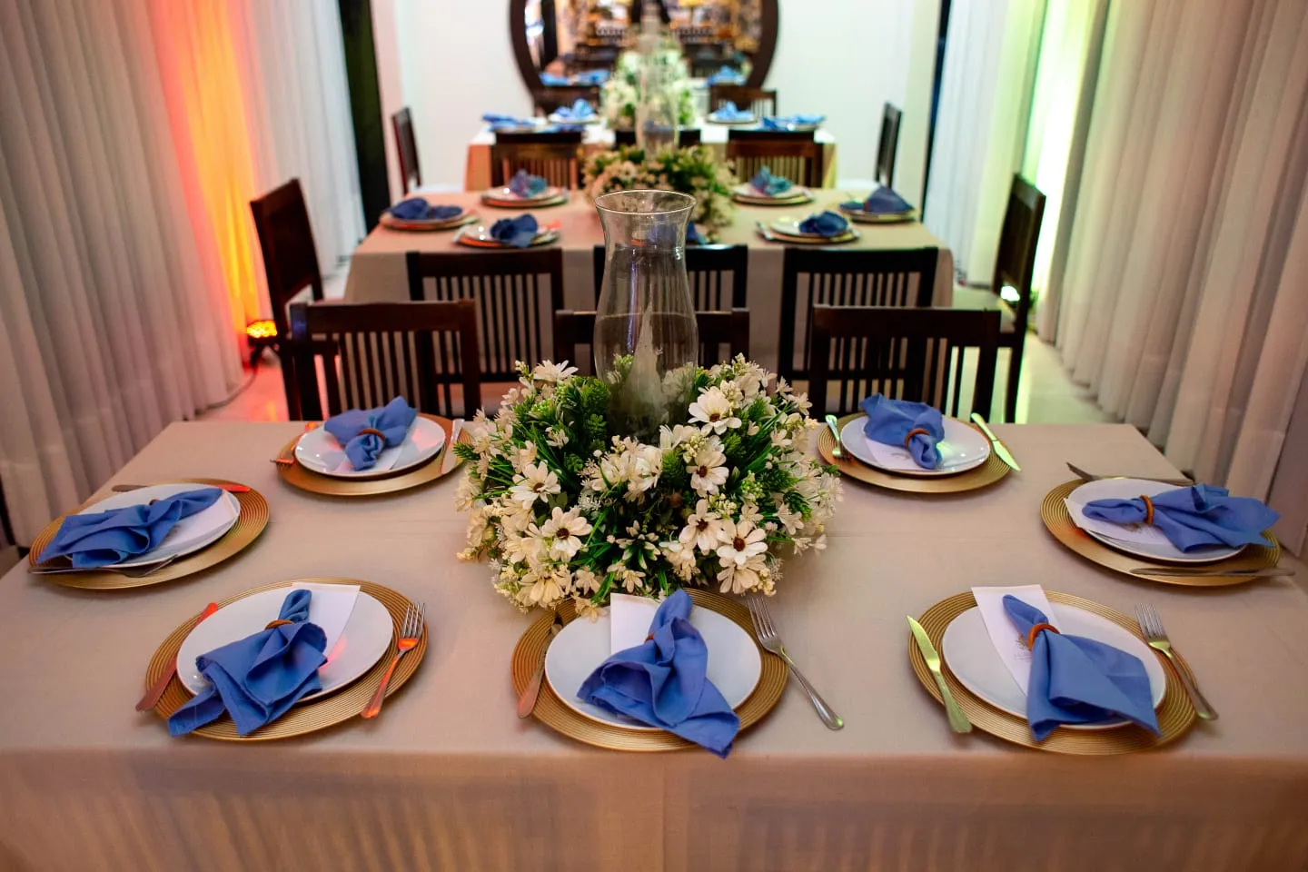 Mesa posta com arranjo de flores brancas, sousplats dourados e guardanapos azuis